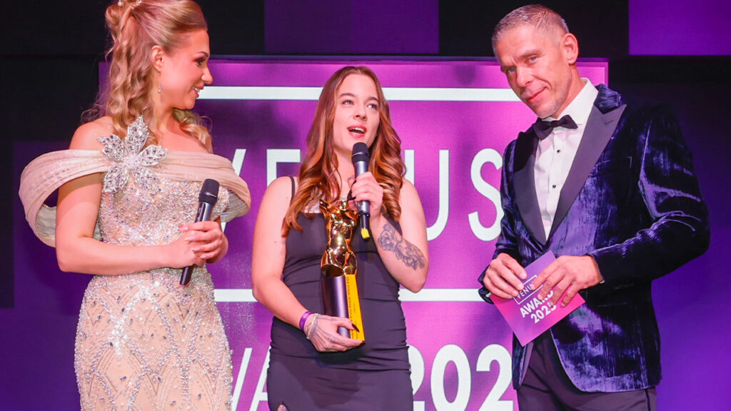 Luna Lynn mit Venus Award 2025 Trophäe hält Mikrofon auf der Bühne zwischen Moderatorin Gabi Gold und Moderator Mick Blue im Wintergarten Varieté Berlin